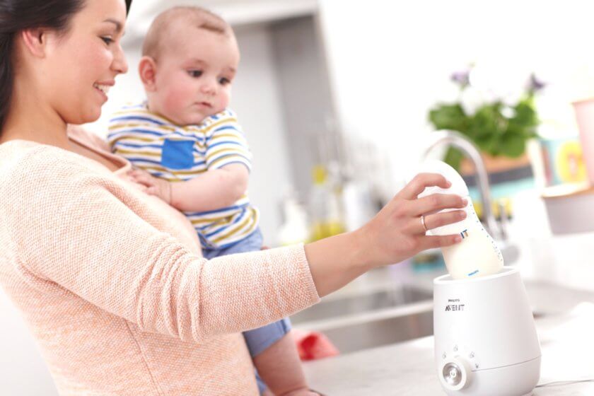 Mutter mit Baby im Arm bereitet einhändig ein Milchfläschchen im Flaschenwärmer zu
